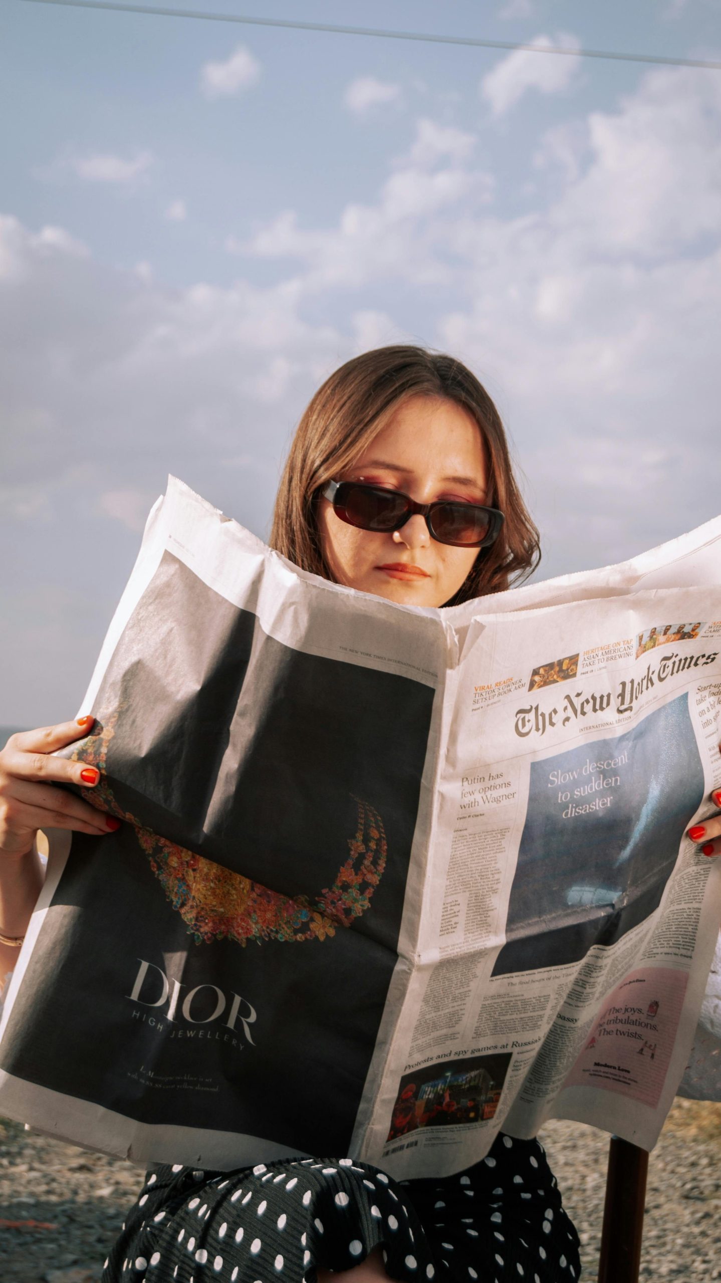Woman enjoys a sunny day reading a newspaper outdoors, wearing sunglasses.
